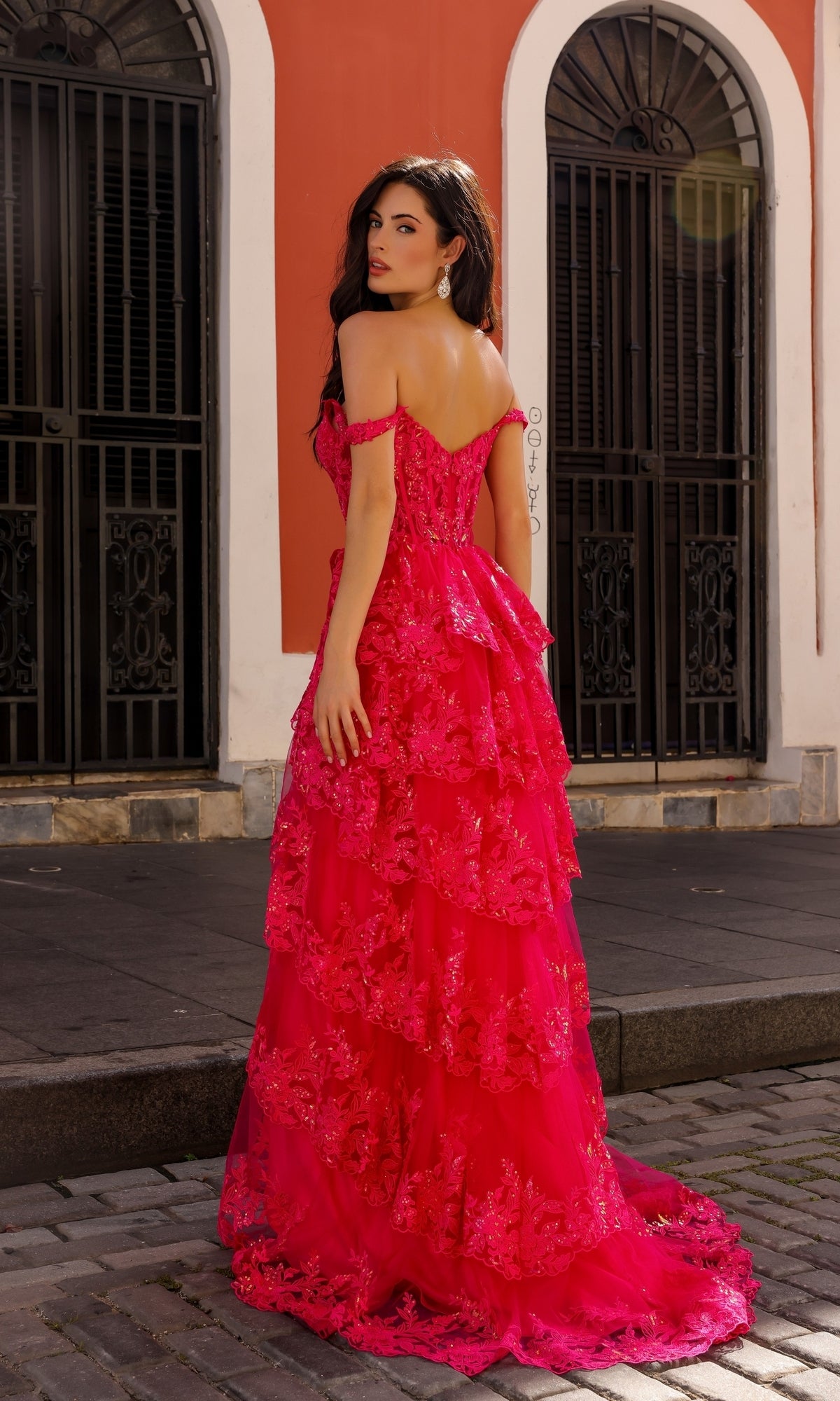 A woman in a vibrant pink Long Lace Ruffled A-Line Prom Dress T1335 stands on a cobblestone street before ornate black double doors and a salmon-colored building—an ideal look for formal occasions.